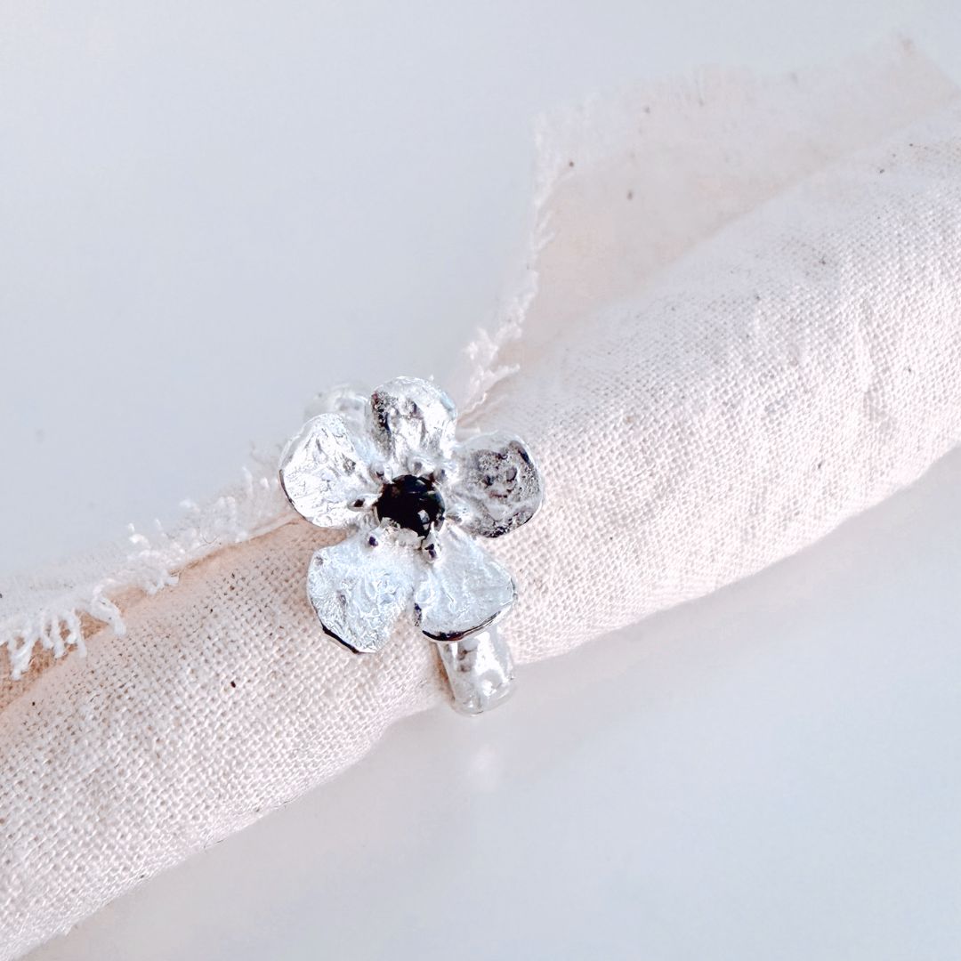 Waiatarua Narrow sterling silver flower ring styled on natural linen, with a dark blue sapphire at the centre.