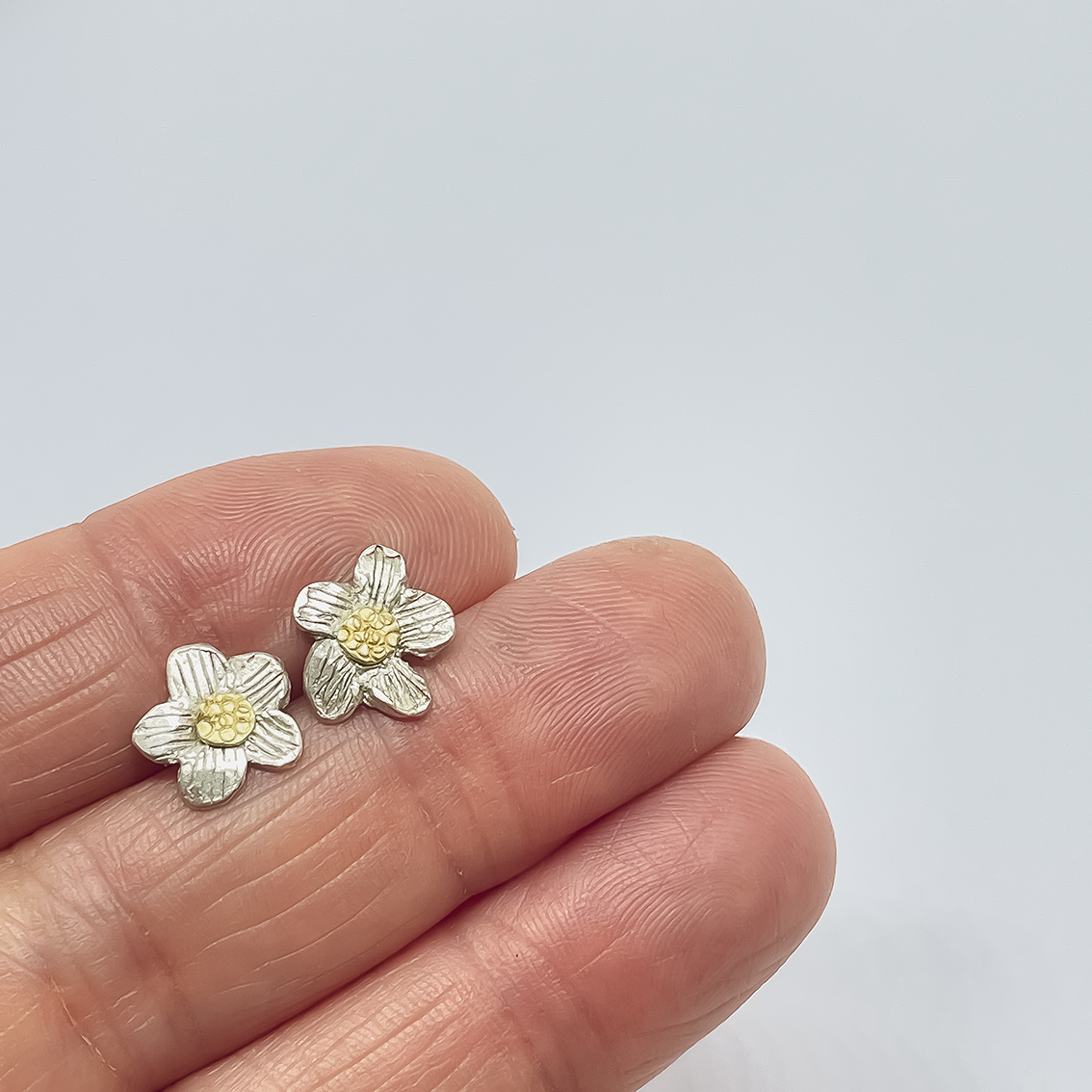 Two small daisy flower studs in Sterling Silver and 18ct yellow Gold centers held between fingers
