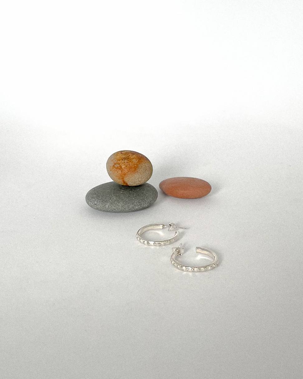 A pair of Textured Narrow Hoop Earrings lying in front of three stones