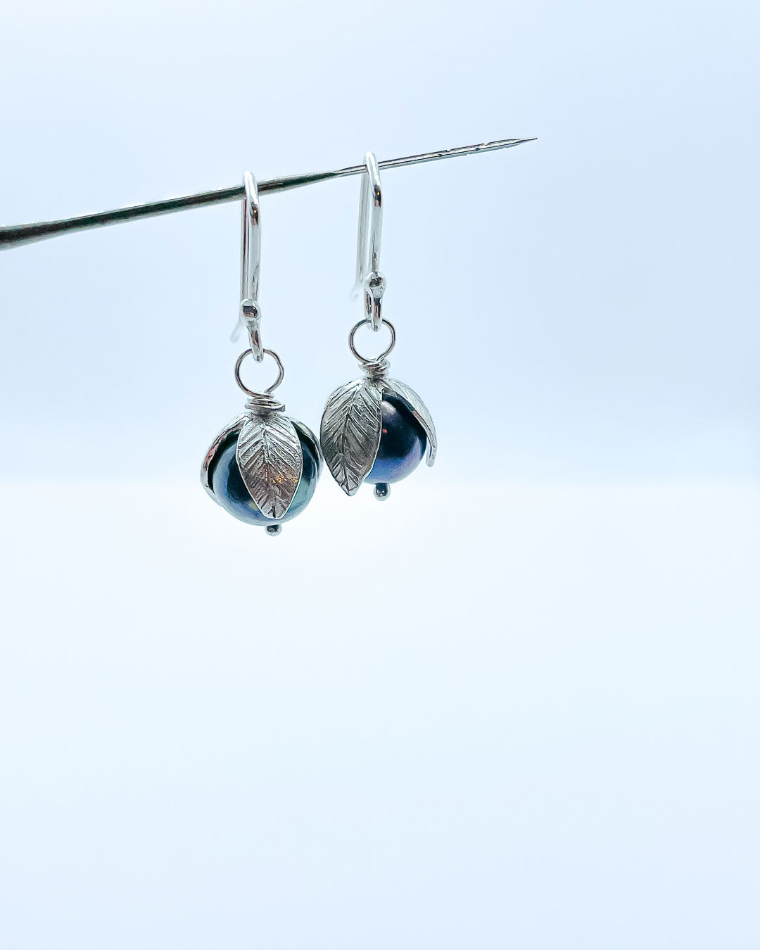 A pair of black pearls topped with Stylised leaves hung from artisan earring hooks hanging from a metal pin