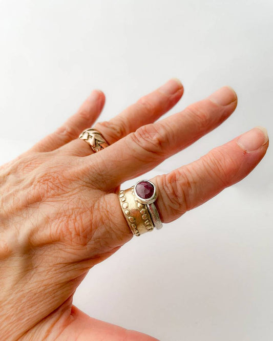A close up of a womans hand that has a wide gold ring and an organic Sterling Silver ring set with a large faceted oval ruby on one of the fingers