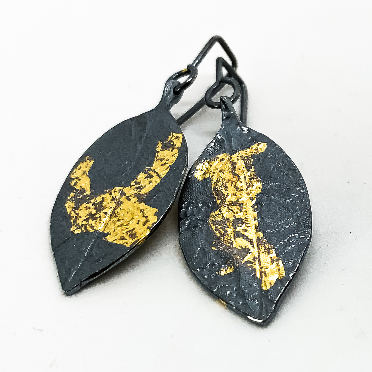 A pair of stylised leaf earrings in Fine Silver that has been oxidised to a deep black with lace embossed into the surface and random patterns of 24ct Gold leaf applied like sunlight dappling the surface