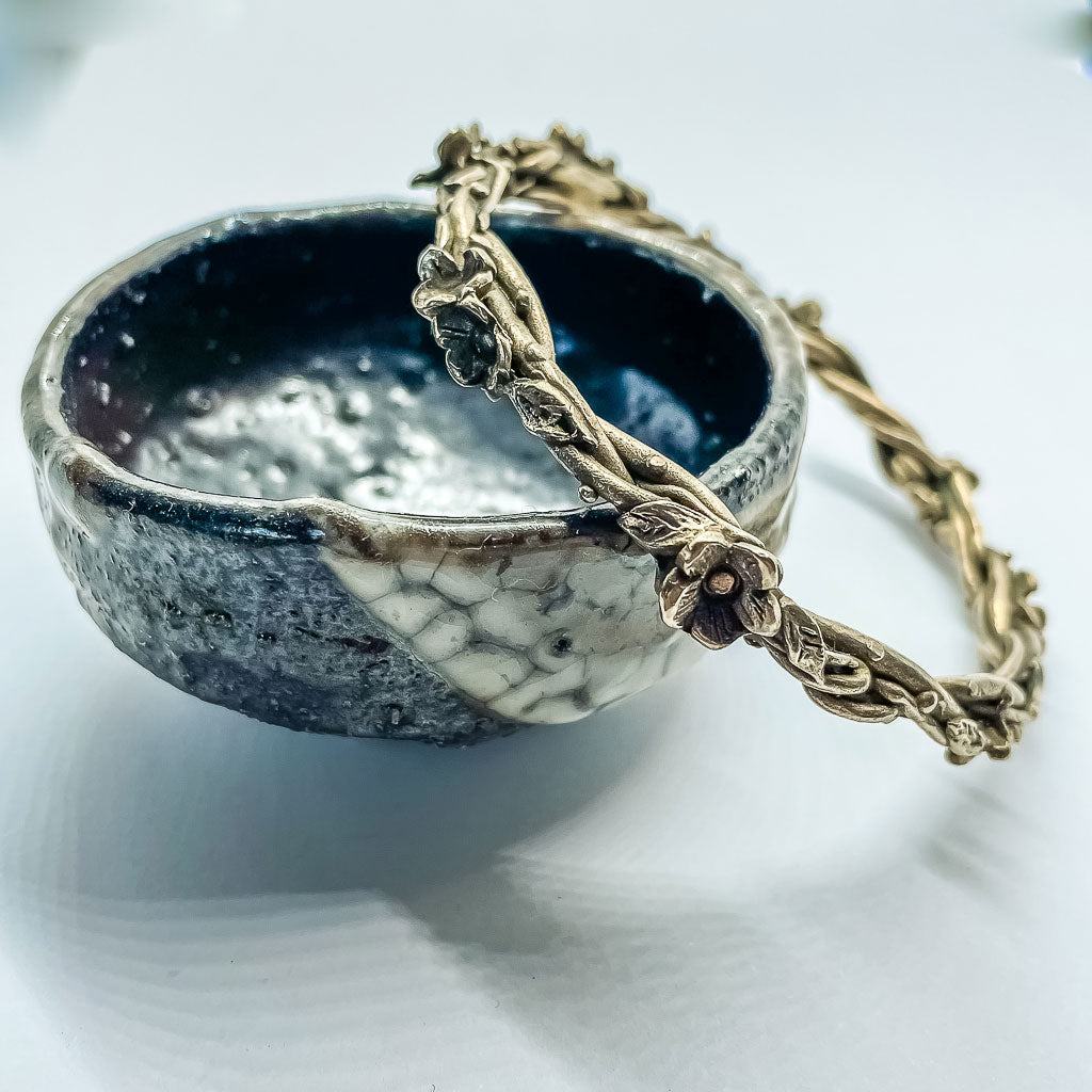 a bronze bangle formed from wrapped vines, leaves and flowers resting against a small artisan pottery bowl