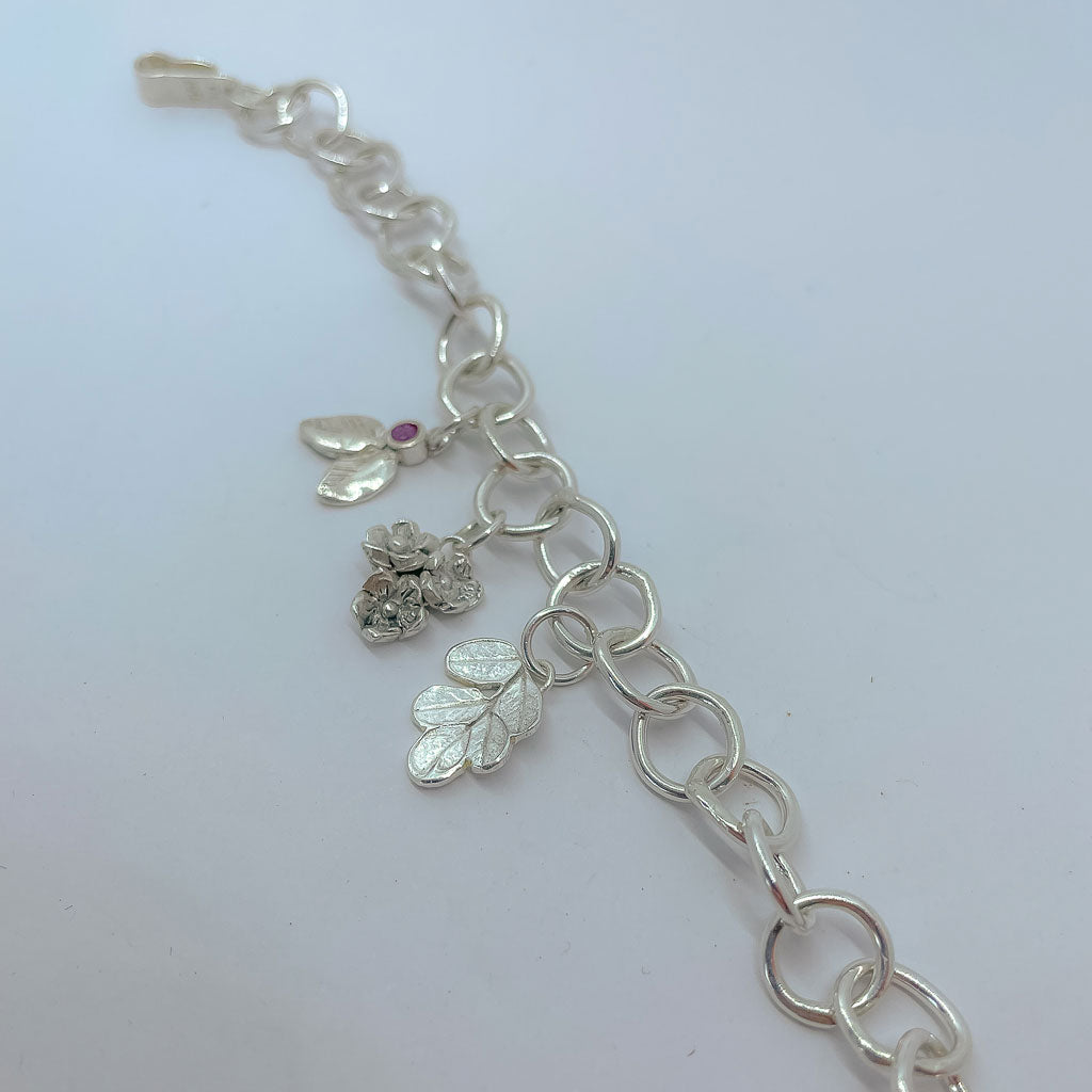 A Silver bracelet made of organic circle links with 3 botanical charms, one a bouquet of flowers, one a Kowhai Leaf print and one textured leaves with a berry set with a ruby