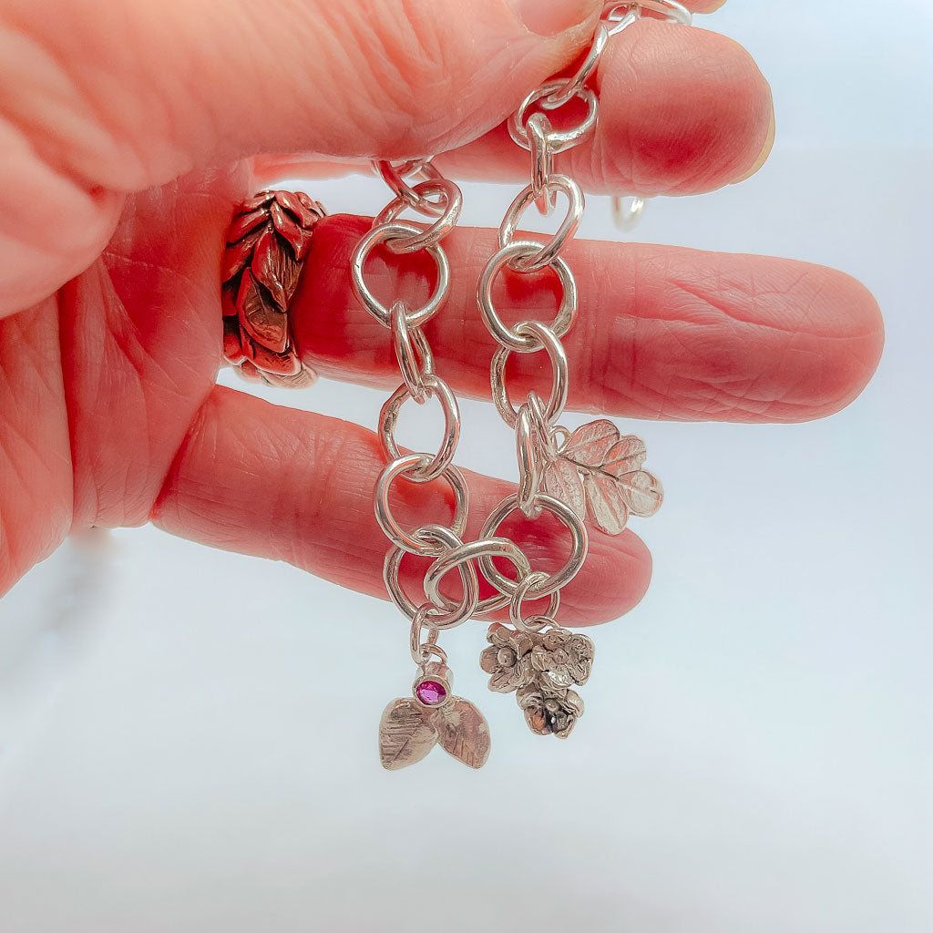 A hand holding a Silver bracelet made of organic circle links with 3 botanical charms, one a bouquet of flowers, one a Kowhai Leaf print and one textured leaves with a berry set with a ruby