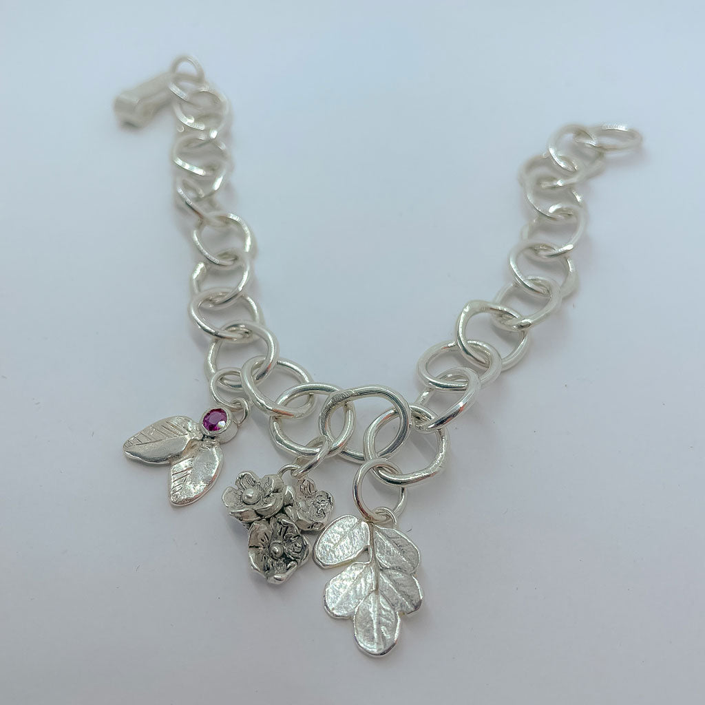 A Silver bracelet made of organic circle links with 3 botanical charms, one a bouquet of flowers, one a Kowhai Leaf print and one textured leaves with a berry set with a ruby
