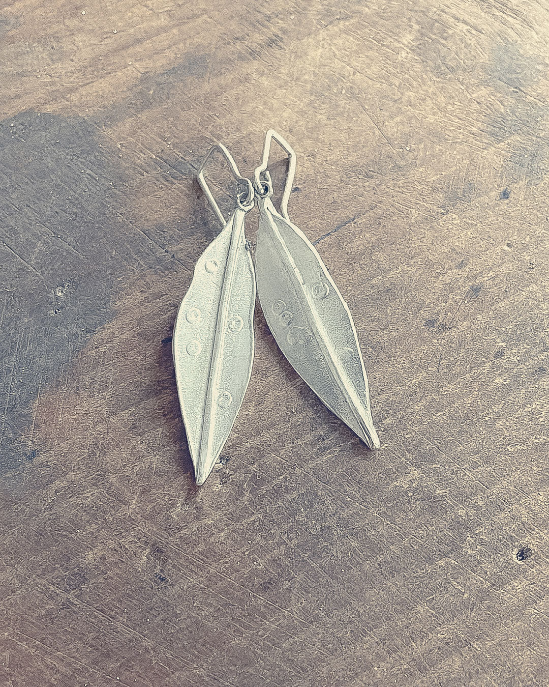 Front profile of a pair of Pohutukawa leaf earrings in Sterling Silver lying on a wood bench